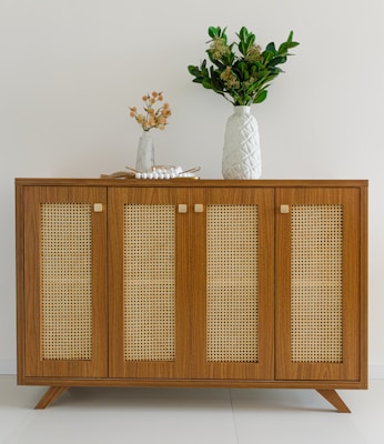 A wooden sideboard with woven rattan panels and modern legs is adorned with a large white vase containing dense green foliage. A smaller white vase with delicate orange flowers and a decorative string of white beads is also present.