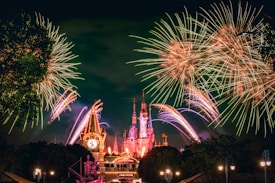 A vibrant display of fireworks illuminates the night sky above a large, ornate castle. The foreground features a clock tower and signage that reads 'Disneyland'. The overall scene is lively and festive, with bursts of colorful lights and the architecture lit in warm hues.