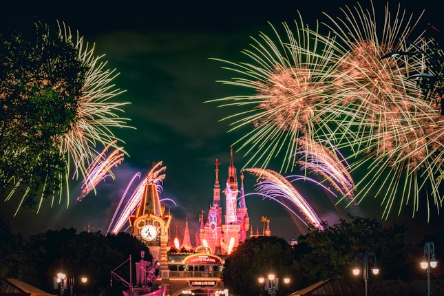A vibrant display of fireworks illuminates the night sky above a large, ornate castle. The foreground features a clock tower and signage that reads 'Disneyland'. The overall scene is lively and festive, with bursts of colorful lights and the architecture lit in warm hues.