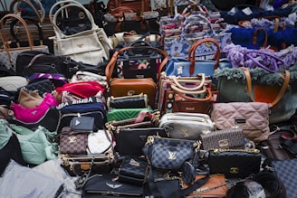 A large assortment of designer handbags displayed closely together. The collection includes various styles, colors, and brands, each featuring distinct textures and patterns. Some bags have visible brand logos and metal hardware accents.