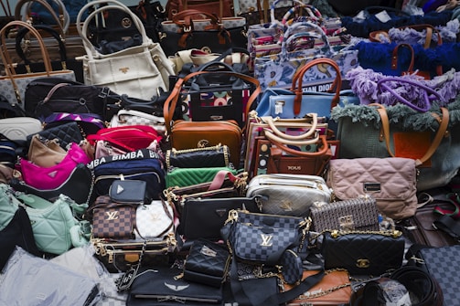 A large assortment of designer handbags displayed closely together. The collection includes various styles, colors, and brands, each featuring distinct textures and patterns. Some bags have visible brand logos and metal hardware accents.