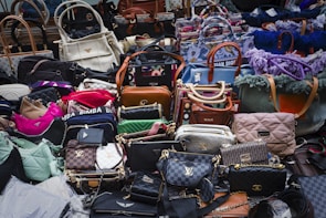 A large assortment of designer handbags displayed closely together. The collection includes various styles, colors, and brands, each featuring distinct textures and patterns. Some bags have visible brand logos and metal hardware accents.