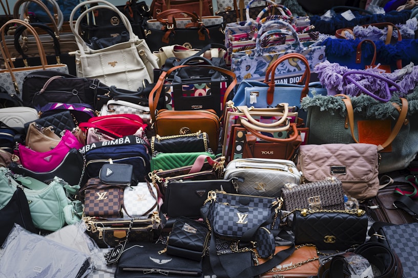 A large assortment of designer handbags displayed closely together. The collection includes various styles, colors, and brands, each featuring distinct textures and patterns. Some bags have visible brand logos and metal hardware accents.