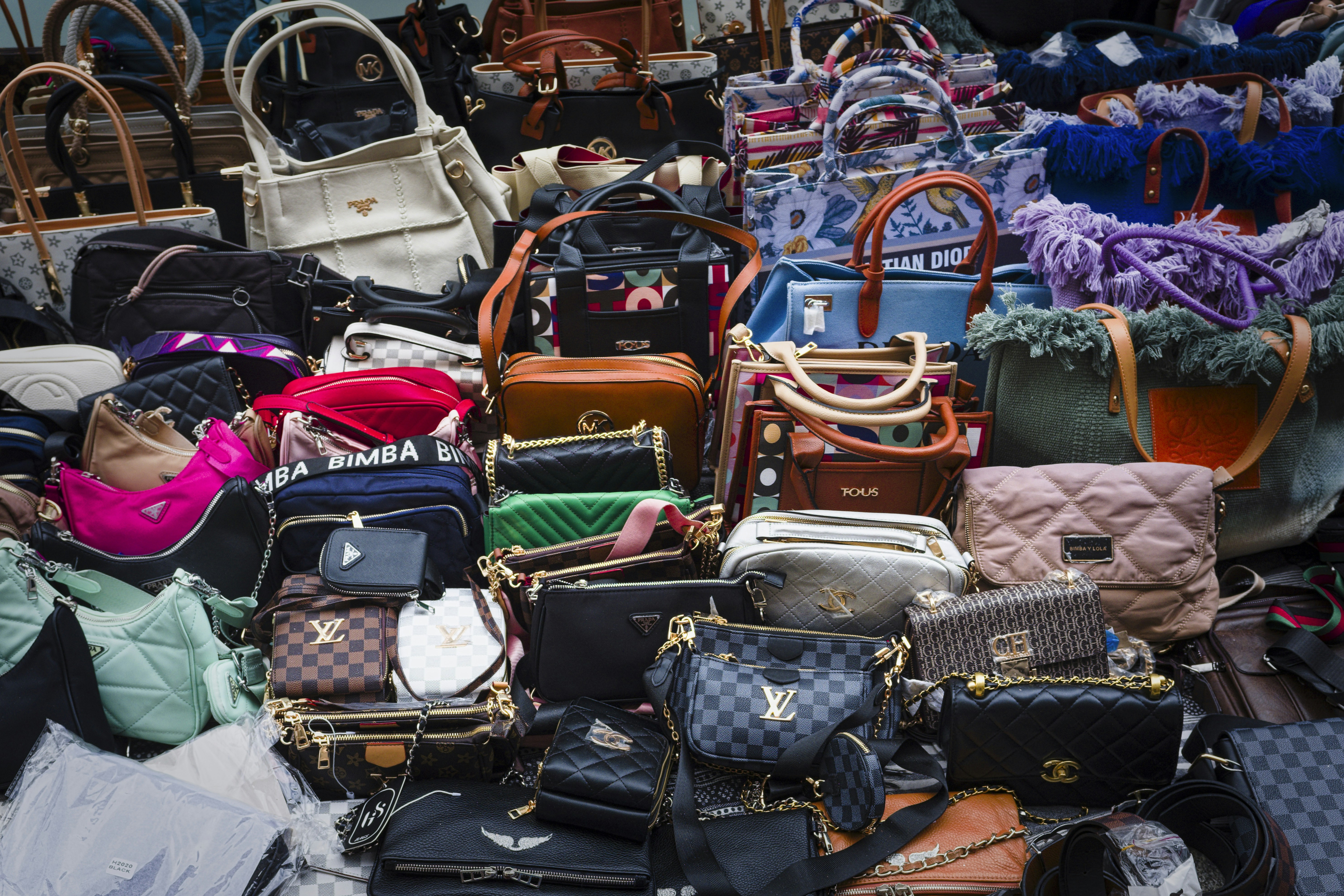 A Large Variety Of Handbags On Display At A Flea Market - A large variety of handbags in various colors, materials, and styles are piled on top of each other at a flea market, with some of the bags displaying brand names.