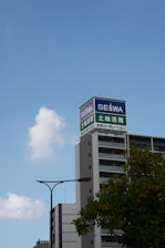 A tall building with a sign on top displaying Japanese characters and the word SEIWA in English is situated against a clear blue sky. Nearby, a modern streetlamp and some greenery can be seen in the foreground.