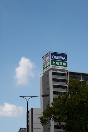 A tall building with a sign on top displaying Japanese characters and the word SEIWA in English is situated against a clear blue sky. Nearby, a modern streetlamp and some greenery can be seen in the foreground.