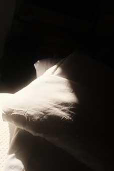 A soft pillow lies partially in sunlight, casting gentle shadows. The light highlights the textures and folds of the fabric, creating a serene and intimate atmosphere.