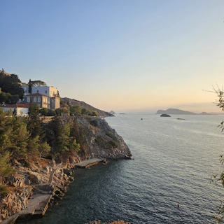 A cozy seaside cottage bathed in warm sunset light, surrounded by lush greenery and a glimpse of the cliffside stairs leading down to the beach.