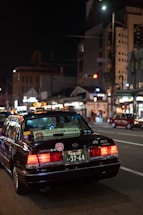 A black taxi with illuminated roof light and various stickers on the rear window is positioned on a city street at night. The license plate is visible and reads '37-64'. The surrounding urban environment includes illuminated buildings and streetlights, contributing to the city's vibrant nightlife ambiance.