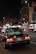 A black taxi with illuminated roof light and various stickers on the rear window is positioned on a city street at night. The license plate is visible and reads '37-64'. The surrounding urban environment includes illuminated buildings and streetlights, contributing to the city's vibrant nightlife ambiance.