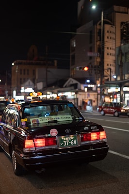 A black taxi with illuminated roof light and various stickers on the rear window is positioned on a city street at night. The license plate is visible and reads '37-64'. The surrounding urban environment includes illuminated buildings and streetlights, contributing to the city's vibrant nightlife ambiance.
