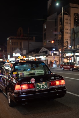 A black taxi with illuminated roof light and various stickers on the rear window is positioned on a city street at night. The license plate is visible and reads '37-64'. The surrounding urban environment includes illuminated buildings and streetlights, contributing to the city's vibrant nightlife ambiance.