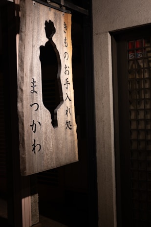 A wooden sign featuring Japanese characters and a cut-out silhouette design, hanging outside on a wall. The sign is illuminated softly, casting shadows against a textured surface.