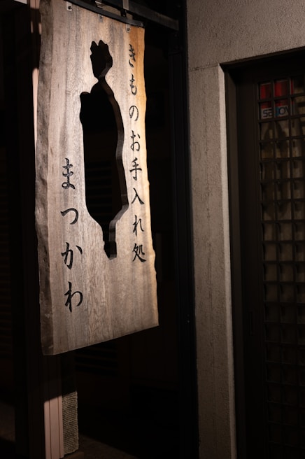 A wooden sign featuring Japanese characters and a cut-out silhouette design, hanging outside on a wall. The sign is illuminated softly, casting shadows against a textured surface.