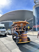 A group of people are seated on a pedal-powered vehicle designed for group tours, resembling a large bicycle with a wooden bar counter and seating. They are traveling on a street in front of a large, modern building with a curved roof and glass façade. The building has a sign that reads 'Bridgestone' and there are other vehicles and pedestrians in the vicinity. An American flag is attached to the top of the vehicle.