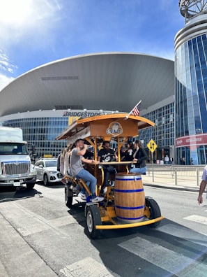 A group of people are seated on a pedal-powered vehicle designed for group tours, resembling a large bicycle with a wooden bar counter and seating. They are traveling on a street in front of a large, modern building with a curved roof and glass façade. The building has a sign that reads 'Bridgestone' and there are other vehicles and pedestrians in the vicinity. An American flag is attached to the top of the vehicle.