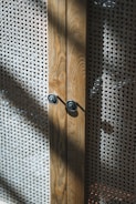 A close-up view of a wooden cabinet with intricate woven mesh panels. The wood has a natural grain, and the cabinet features two circular metallic knobs. Sunlight casts shadows across the textured surface of the mesh, creating a play of light and shadow.