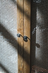 A close-up view of a wooden cabinet with intricate woven mesh panels. The wood has a natural grain, and the cabinet features two circular metallic knobs. Sunlight casts shadows across the textured surface of the mesh, creating a play of light and shadow.