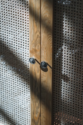 A close-up view of a wooden cabinet with intricate woven mesh panels. The wood has a natural grain, and the cabinet features two circular metallic knobs. Sunlight casts shadows across the textured surface of the mesh, creating a play of light and shadow.