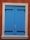 A bright blue wooden shutter with black metal hinges is tightly closed against an orange-brown stucco wall. The frame surrounding the shutter is a light beige stone, creating a contrast with the wall and shutters. A small piece of grass or straw is wedged between the shutters' doors.