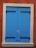 A bright blue wooden shutter with black metal hinges is tightly closed against an orange-brown stucco wall. The frame surrounding the shutter is a light beige stone, creating a contrast with the wall and shutters. A small piece of grass or straw is wedged between the shutters' doors.
