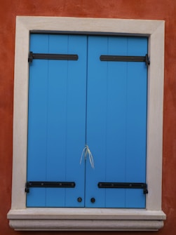 A bright blue wooden shutter with black metal hinges is tightly closed against an orange-brown stucco wall. The frame surrounding the shutter is a light beige stone, creating a contrast with the wall and shutters. A small piece of grass or straw is wedged between the shutters' doors.