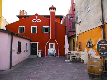 A colorful and quaint alley featuring vibrant buildings with red, pink, and orange facades. The central red building has green shuttered windows and white decorative details. An outdoor seating area with chairs and tables is on the right, next to a rustic wooden barrel. Artistic wall decorations and a vintage-style light fixture add charm to the scene.