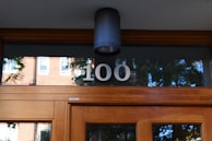 A modern wooden entrance door featuring a frosted glass window with the number 100 displayed. Above the door is a sleek black wall-mounted light fixture. The reflection of trees and a brick building can be seen in the glass window.