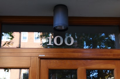 A modern wooden entrance door featuring a frosted glass window with the number 100 displayed. Above the door is a sleek black wall-mounted light fixture. The reflection of trees and a brick building can be seen in the glass window.