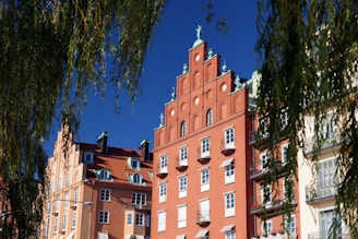 A group of colorful, historical buildings with ornate architectural details. The red brick building in the center features gables, decorative sculptures, and multiple windows, while the adjacent structures are in warm tones with similar architectural elements. Overhanging tree branches frame the scene against a clear blue sky.