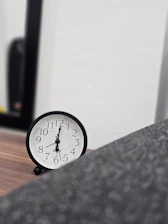A black analog clock with white face and black numbers sitting on a wooden surface. The background includes a partial view of a dark-framed mirror and a blurred gray object in the foreground.