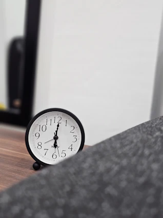 A black analog clock with white face and black numbers sitting on a wooden surface. The background includes a partial view of a dark-framed mirror and a blurred gray object in the foreground.