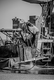 A construction worker is operating heavy machinery, possibly involved in roadwork or industrial activity. The person is wearing protective clothing, including a vest and helmet, and is bent over the machine, focused on the task. The machinery is complex, with various levers, hoses, and metal surfaces, and appears to be engaged in a process involving liquid or water on the ground.