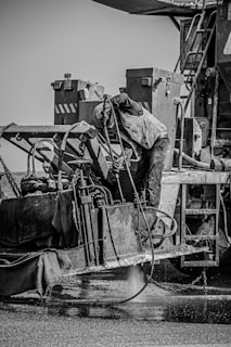 A construction worker is operating heavy machinery, possibly involved in roadwork or industrial activity. The person is wearing protective clothing, including a vest and helmet, and is bent over the machine, focused on the task. The machinery is complex, with various levers, hoses, and metal surfaces, and appears to be engaged in a process involving liquid or water on the ground.