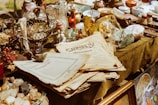 A variety of antique and vintage items are spread across a table. These include old newspapers, brass and glass bottles, ornate figurines, a wire basket filled with spherical objects, and large crystals or minerals. The scene conveys a sense of history and nostalgia.