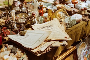 A variety of antique and vintage items are spread across a table. These include old newspapers, brass and glass bottles, ornate figurines, a wire basket filled with spherical objects, and large crystals or minerals. The scene conveys a sense of history and nostalgia.