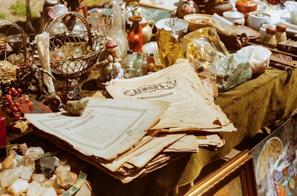 A variety of antique and vintage items are spread across a table. These include old newspapers, brass and glass bottles, ornate figurines, a wire basket filled with spherical objects, and large crystals or minerals. The scene conveys a sense of history and nostalgia.