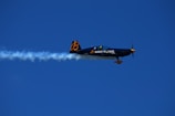 An aerobatic airplane is flying through a clear blue sky, leaving a trail of white smoke behind. The plane is decorated with the logo and branding of Breitling in yellow and white.