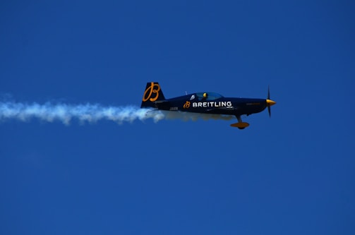 An aerobatic airplane is flying through a clear blue sky, leaving a trail of white smoke behind. The plane is decorated with the logo and branding of Breitling in yellow and white.