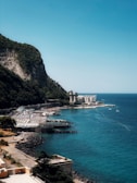 A scenic coastal area with a rocky cliff covered in greenery on the left. Below the cliff, there is a shoreline with clear, turquoise water and a small resort area featuring rows of sun loungers and umbrellas. A large, white building with a classical architectural design is seen in the distance, near the edge of the water. A few boats are visible in the sea.