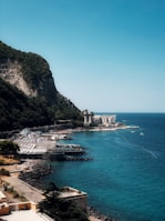 A scenic coastal area with a rocky cliff covered in greenery on the left. Below the cliff, there is a shoreline with clear, turquoise water and a small resort area featuring rows of sun loungers and umbrellas. A large, white building with a classical architectural design is seen in the distance, near the edge of the water. A few boats are visible in the sea.
