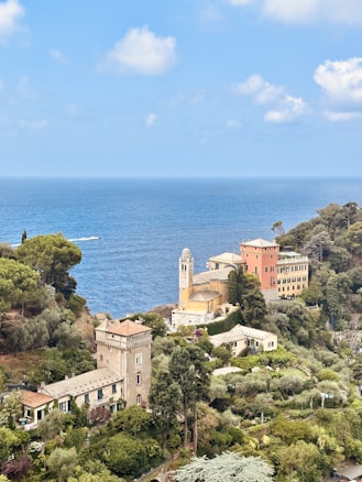 A picturesque coastal village is nestled against the backdrop of a serene, azure sea. The landscape features charming buildings with Mediterranean architecture, surrounded by lush greenery and trees. A prominent church with a clock tower stands out among the buildings, adding a historical element to the scene. The sky is partly cloudy, enhancing the tranquil ambiance.
