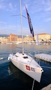A white sailboat docked at a marina with the registration number 810. The boat features a tall mast with banners, surrounded by calm water. In the background, several historical buildings line the waterfront, under a clear blue sky.