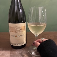A bottle of Chardonnay with the label 'Vine in Flames' alongside a glass of white wine. A hand with rings is holding the glass. The background is a muted green, and the surface is a dark wood table.