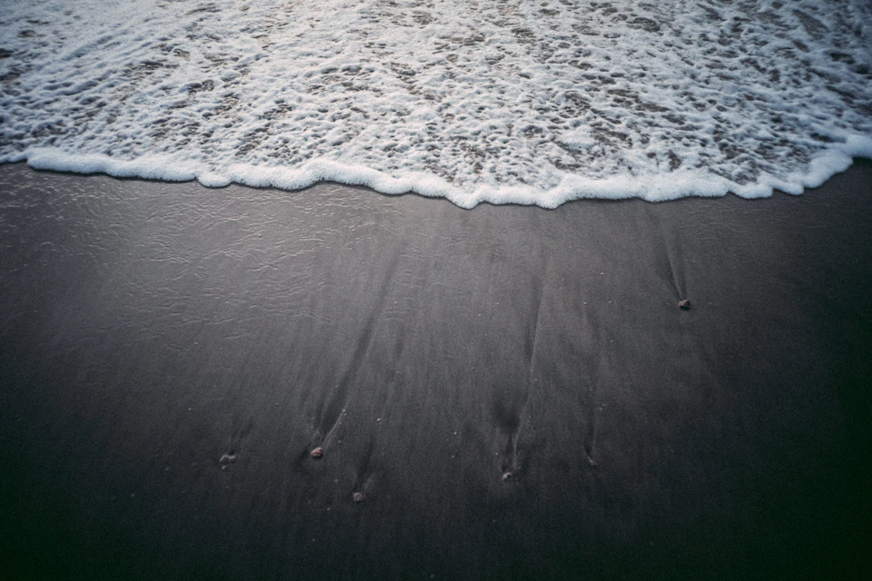 Serene black sand beach with foamy light blue water and surrounding landscape from above