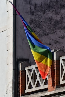 A rainbow flag is hanging from a building with a textured, weathered gray wall. The flag, displaying vibrant colors, is attached to a metal pole extending from a brick and stone balcony adorned with geometric patterns.