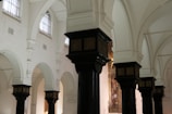 The image depicts the interior of a historic building with ornate, dark wooden columns topped with decorative capitals. The walls are painted white, and there are high, arched windows allowing soft natural light to illuminate the space. The architecture features a series of arches and detailed moldings.