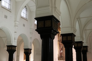 The image depicts the interior of a historic building with ornate, dark wooden columns topped with decorative capitals. The walls are painted white, and there are high, arched windows allowing soft natural light to illuminate the space. The architecture features a series of arches and detailed moldings.
