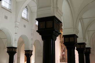 The image depicts the interior of a historic building with ornate, dark wooden columns topped with decorative capitals. The walls are painted white, and there are high, arched windows allowing soft natural light to illuminate the space. The architecture features a series of arches and detailed moldings.