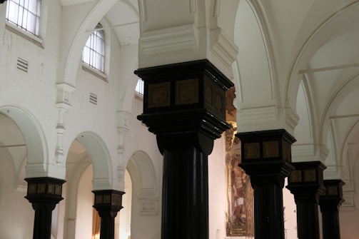 The image depicts the interior of a historic building with ornate, dark wooden columns topped with decorative capitals. The walls are painted white, and there are high, arched windows allowing soft natural light to illuminate the space. The architecture features a series of arches and detailed moldings.